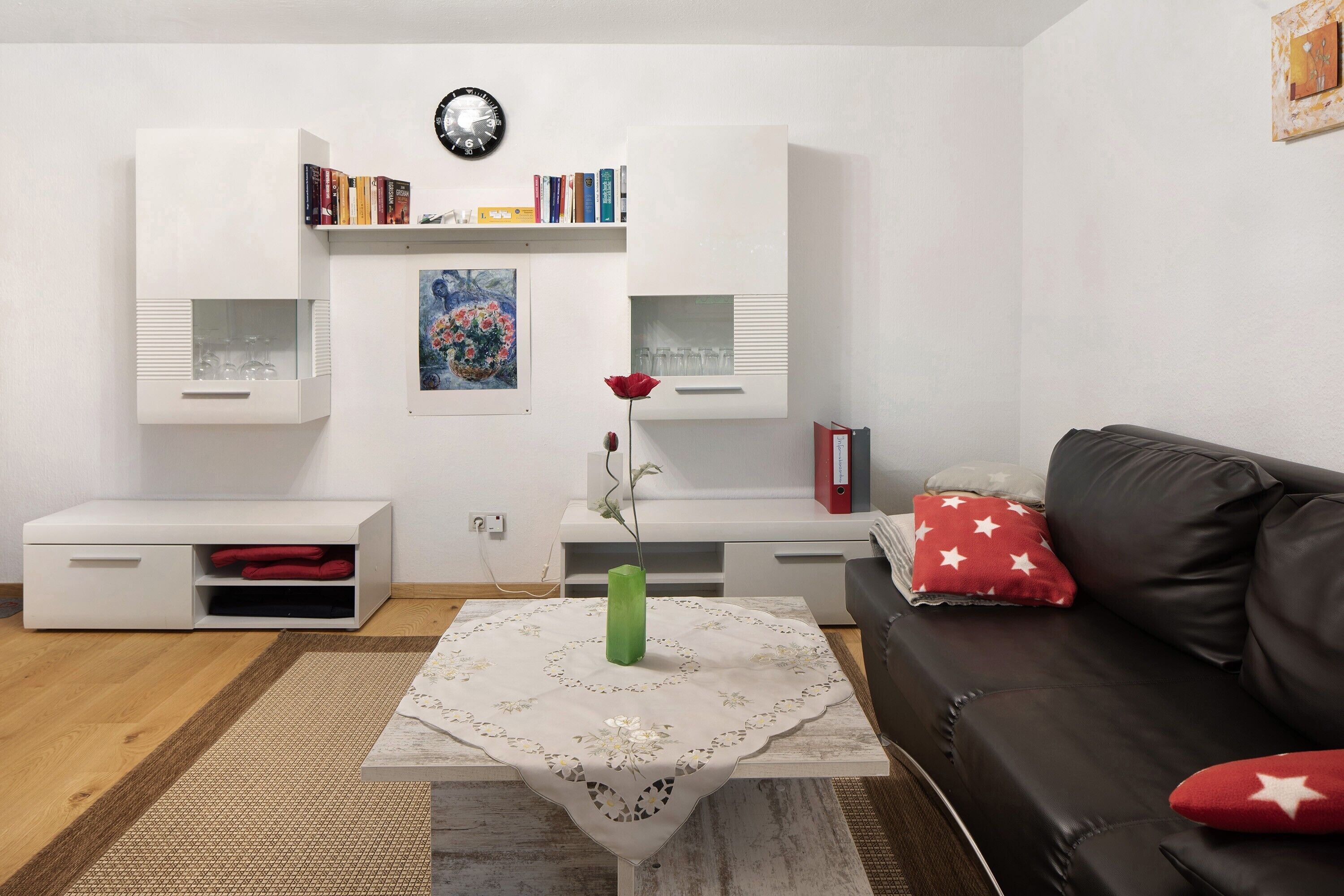 Ein Wohnzimmer mit einer Couch, einem Couchtisch mit einer Vase, einem Bücherregal und einer Uhr an der Wand.