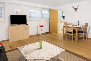 Una habitación con una televisión sobre un mueble de madera, una mesa de comedor con sillas, un florero con una flor y una ventana con persianas.
