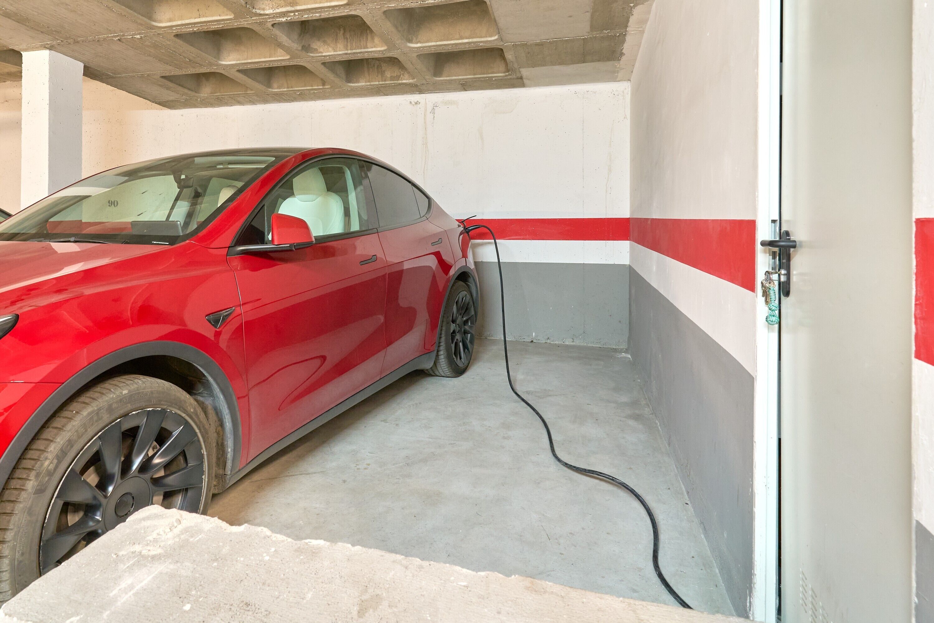 Un coche rojo estacionado en un garaje con suelo de concreto y una puerta con una franja roja y blanca.