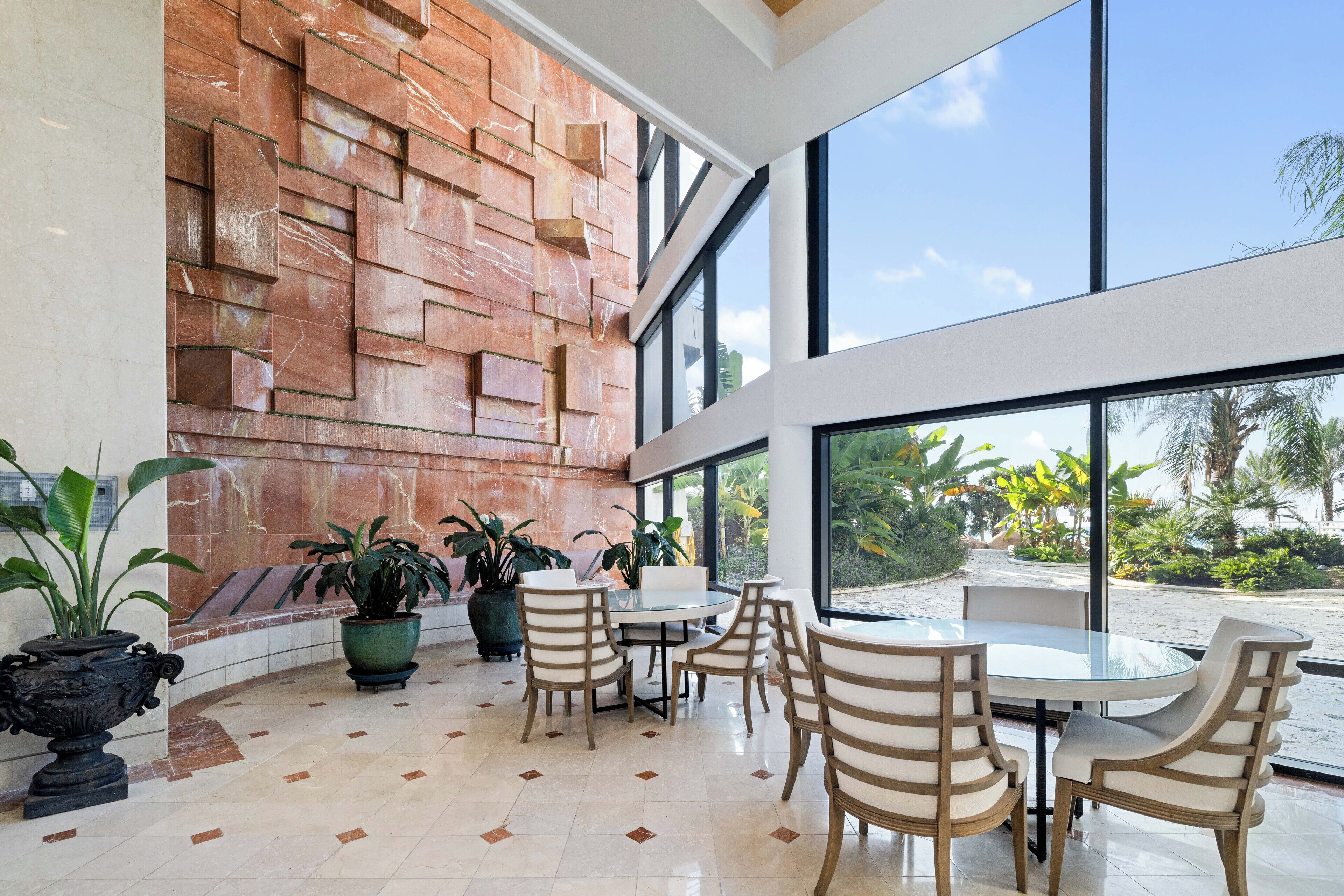 Un espace intérieur vaste et lumineux, doté d’une grande fenêtre donnant sur une plage, avec un sol carrelé, plusieurs plantes en pot et une table ronde entourée de chaises.