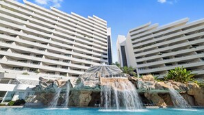 Un hotel moderno con varios balcones, una piscina con cascada y un cielo azul despejado.