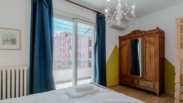 A bedroom with a bed, a wooden wardrobe, a chandelier, and a view of buildings through a window. - GuestHost - Bright Apartment Lingotto Torino (Turin)