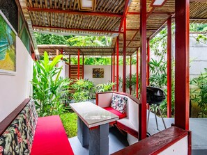 Un espace extérieur aménagé, abrité et doté d’une table et d’un banc, entouré d’une végétation luxuriante et surmonté d’une pergola en bois.