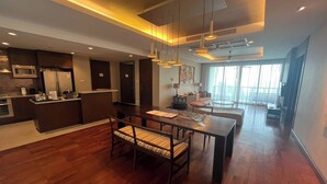 A spacious living area with a wooden dining table, a kitchen with modern appliances, and a large window providing natural light. - A Shasa Vela Condominium (Koh Samui)