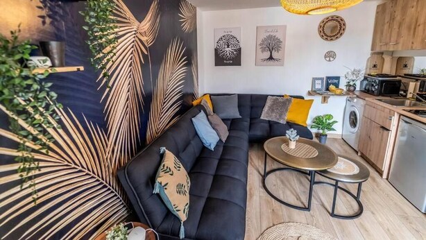 A modern living room with a black sofa, a round coffee table, and a kitchen area with wooden cabinets and appliances.