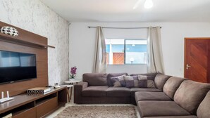 A living room with a large flat-screen TV, a brown sofa, a small side table with a potted plant, and a window with curtains. - Casa em Itanhaém Pé na Areia com Piscina (Itanhaém)