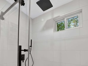 Un baño moderno con cabina de ducha de vidrio, paredes con azulejos blancos y una ventana con vistas a árboles.
