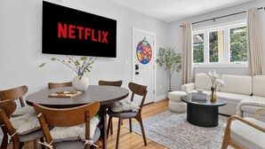 Moderni olohuone, jossa on ruokapöytä, tuoleja, sohva, sohvapöytä ja seinälle asennettu televisio, jonka näytöllä näkyy teksti ”NETFLIX”.