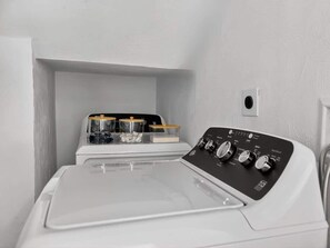 Un appareil compact combinant lave-linge et sèche-linge, doté d’un panneau de commande, d’une petite étagère avec des récipients et d’une prise de courant murale.