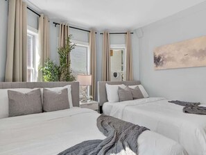 A hotel room with two beds, a nightstand, a lamp, a plant, and a painting on the wall.
