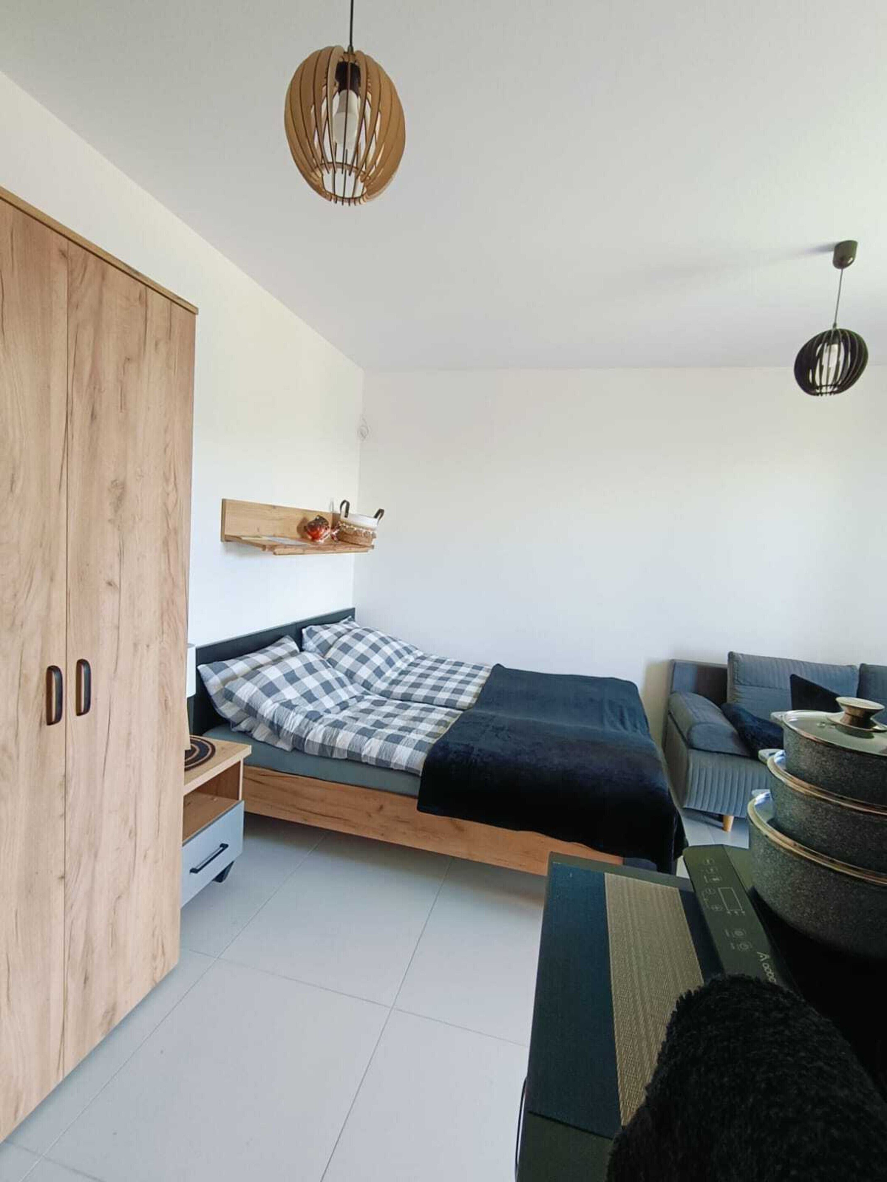 Ein modernes Schlafzimmer mit einem Holzschrank, einem Bett mit kariertem Überwurf, einem grauen Sofa und einer Pendelleuchte.