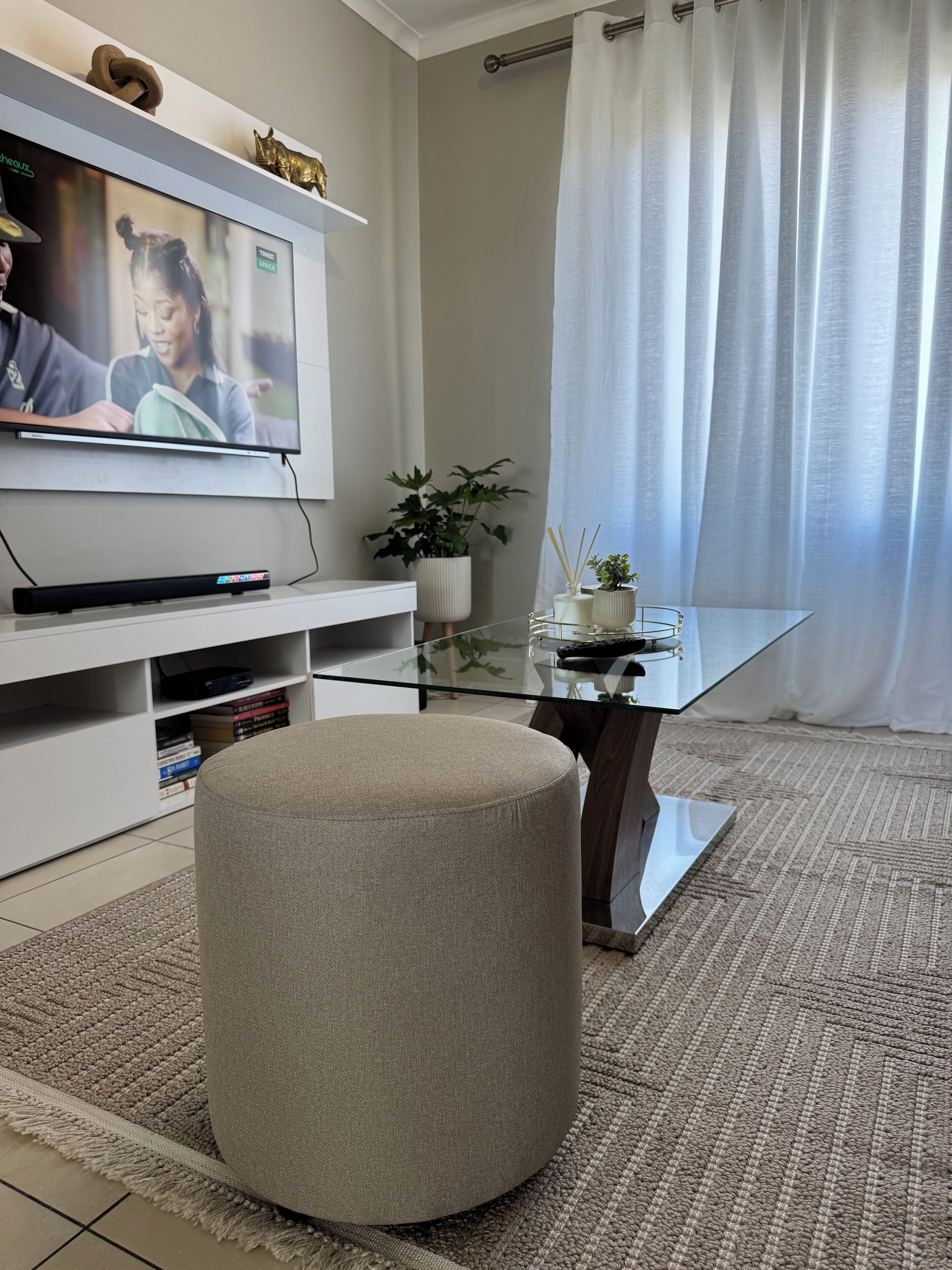A living room with a flat-screen TV, a glass coffee table, a round ottoman, a white entertainment unit, and a potted plant.