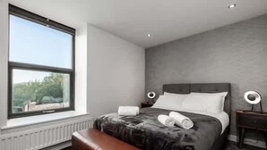A bedroom with a large window, a bed with a dark blanket, a bedside table with a lamp, and a bench. - Victoria Home (Hyde)