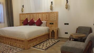 Un dormitorio con una cama de madera, almohadas rojas, una mesita de noche de madera, un sillón gris y una mesa redonda de madera.