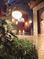 Un patio con un gran lámpara decorativa sobre una zona de estar, rodeado de plantas en macetas y una pared de ladrillo.