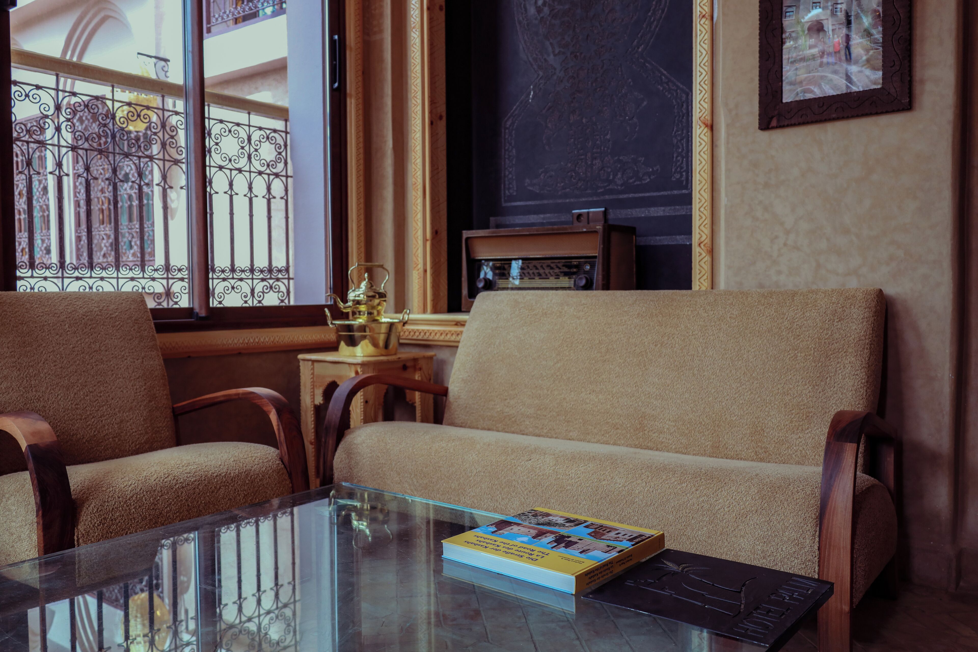 Une pièce comprenant un canapé, des fauteuils, une table en verre, un livre, une radio et une vue sur l’extérieur, avec un balcon accessible par une fenêtre.