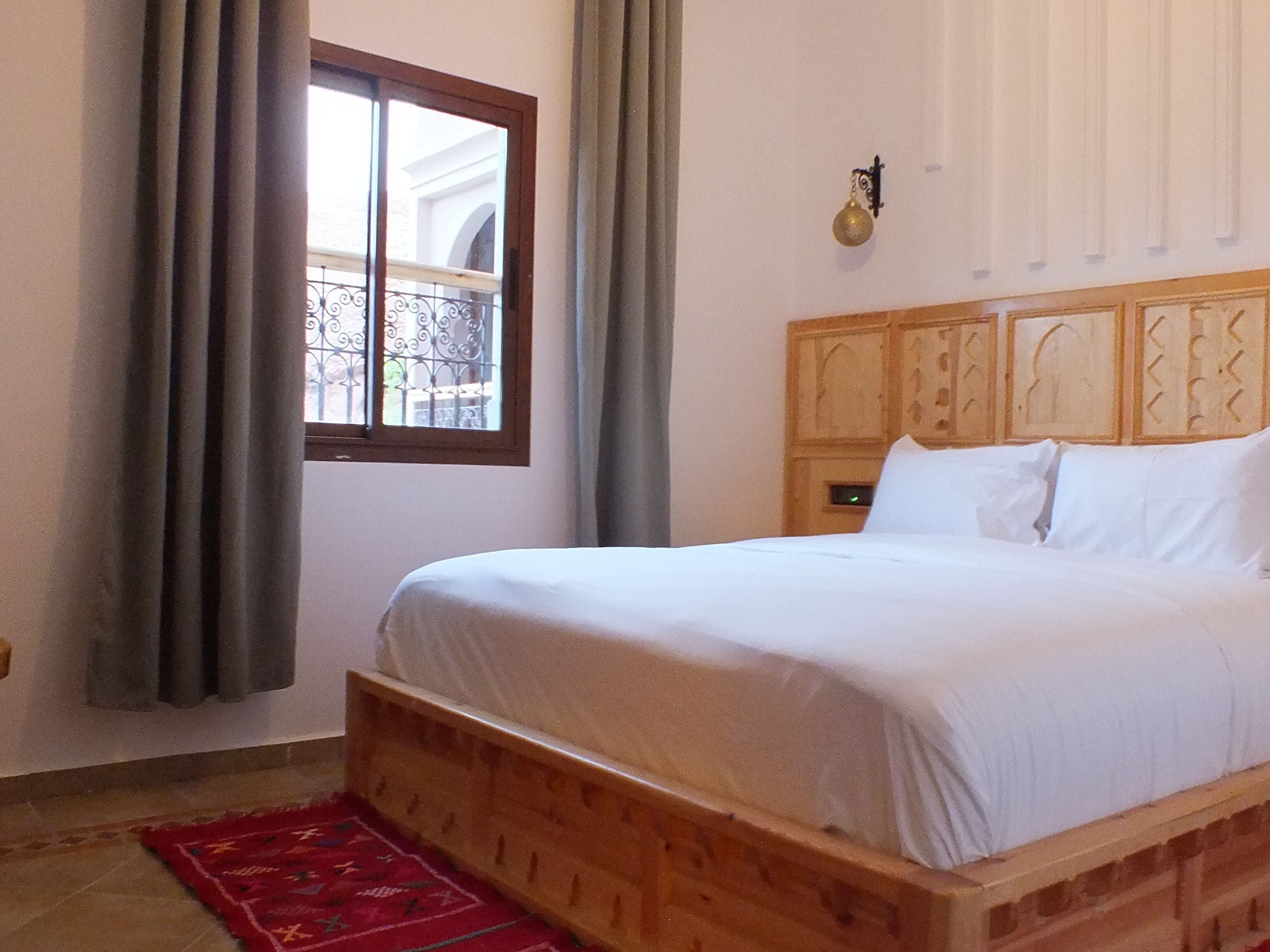 A room with a wooden bed, a red patterned rug, a window with curtains, and a wall-mounted light fixture.