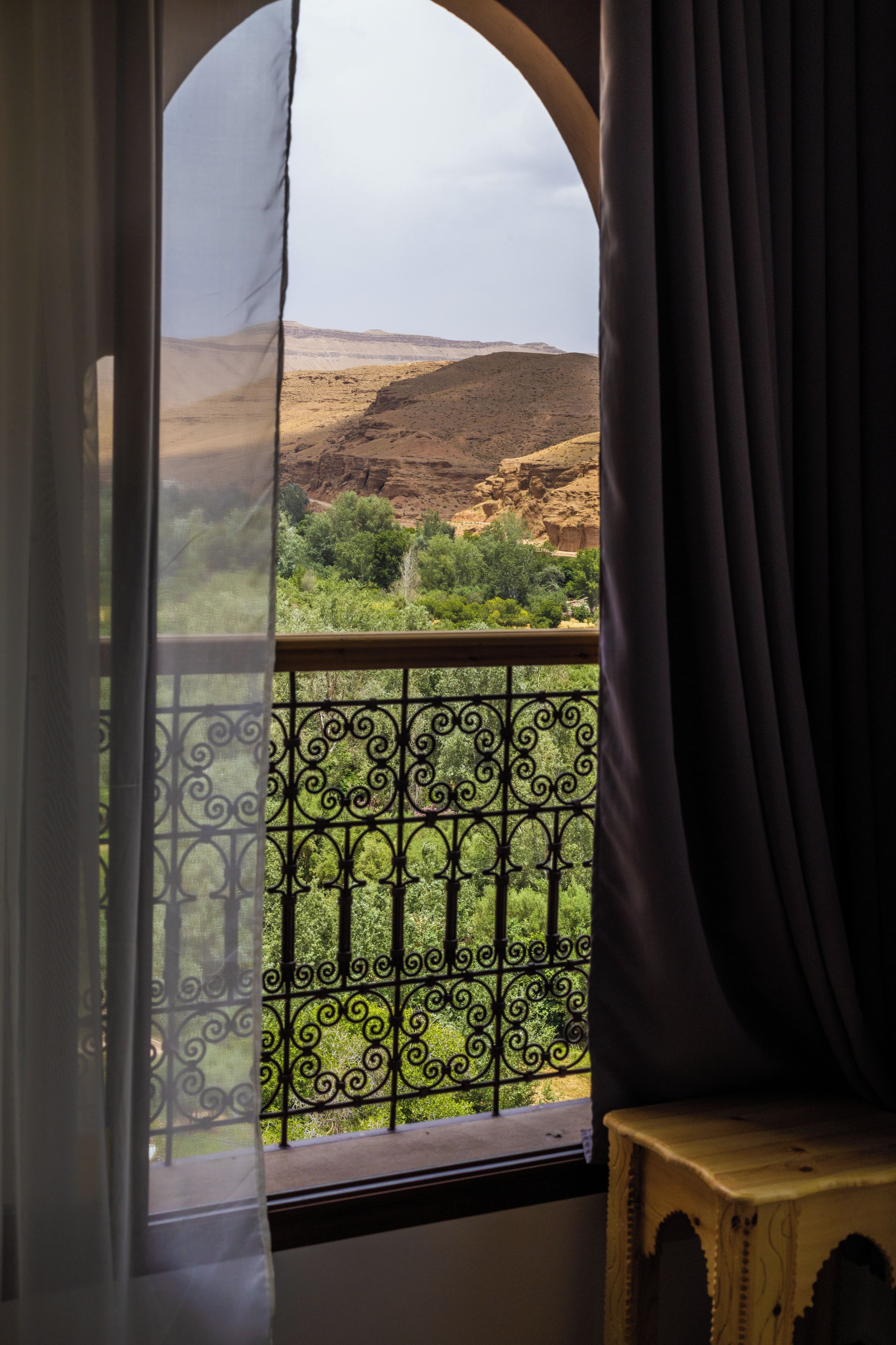 Une fenêtre donnant sur un paysage montagneux, un balcon orné d’une balustrade en fer forgé et un rideau tiré partiellement sur le côté.