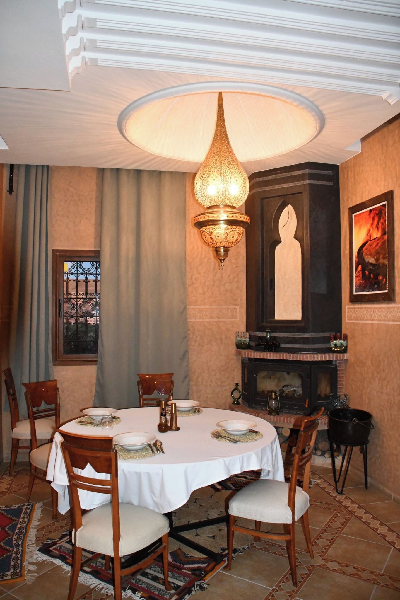 A dining room with a round table set for four, a decorative wall-mounted light fixture, a fireplace, and a window with curtains.