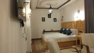 A bedroom with a wooden bed, a chair, a small table, and a wall-mounted TV.