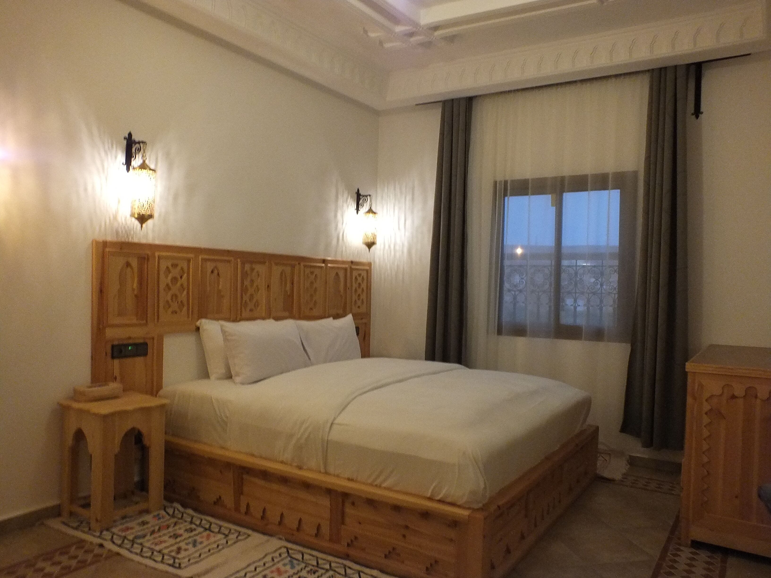 A bedroom with a wooden bed, two wall-mounted lights, a nightstand, a small cabinet, and a window with curtains.