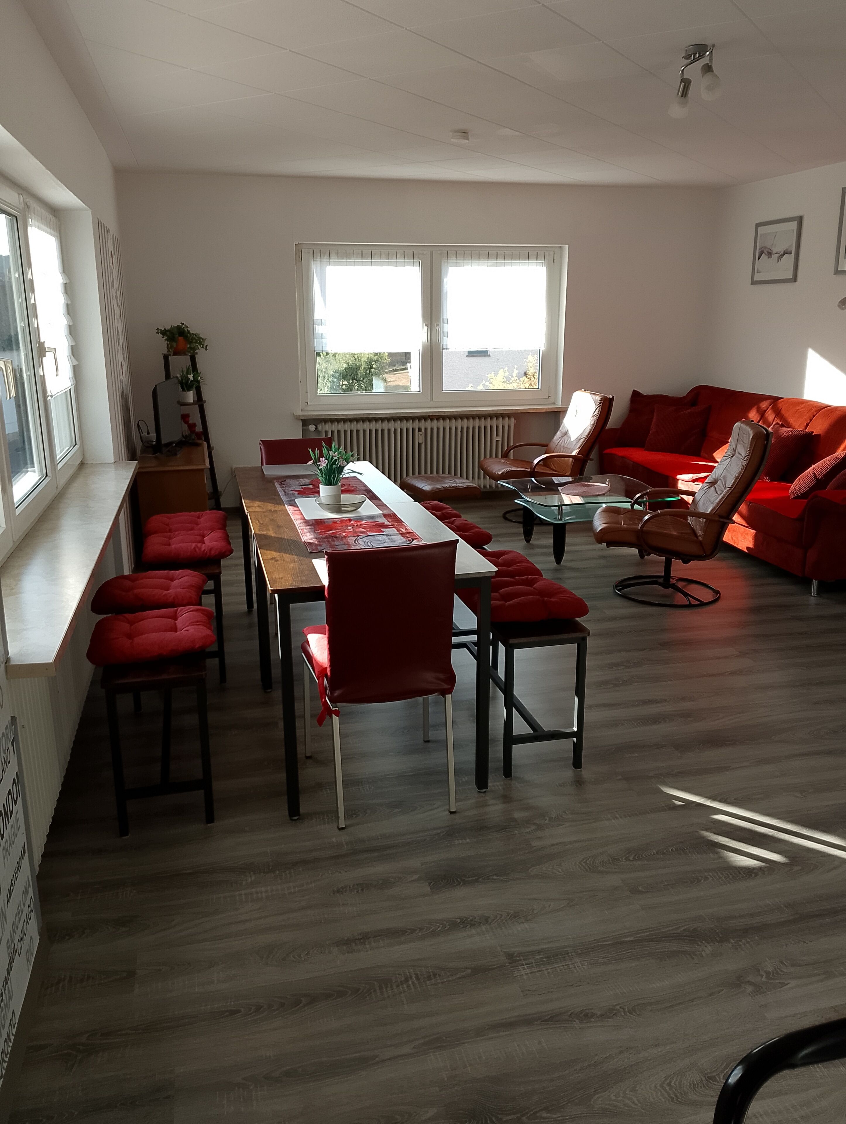 Uma sala de estar moderna com área de jantar, piso de madeira, mobiliário vermelho e grandes janelas com persianas.
