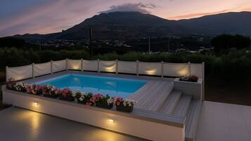 A modern outdoor pool with a wooden deck and white seating, surrounded by flower planters, set against a mountainous backdrop.