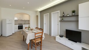 A modern kitchen with white cabinets, a dining table set for four, and a flat-screen TV on a white stand. - Villa Corallo in Scopello (Castellammare del Golfo)
