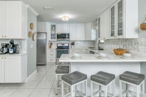 Et moderne køkken med hvide skabe, en central ø med barstole, rustfrie stål hvidevarer og et backsplash med fiskebensmønstret fliser.