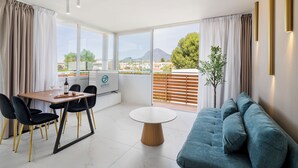Una sala de estar moderna con un sofá, una mesa de comedor y sillas. Hay un televisor sobre un mueble bajo, una planta en maceta, y vistas a la ciudad y las montañas a través de amplias ventanas.