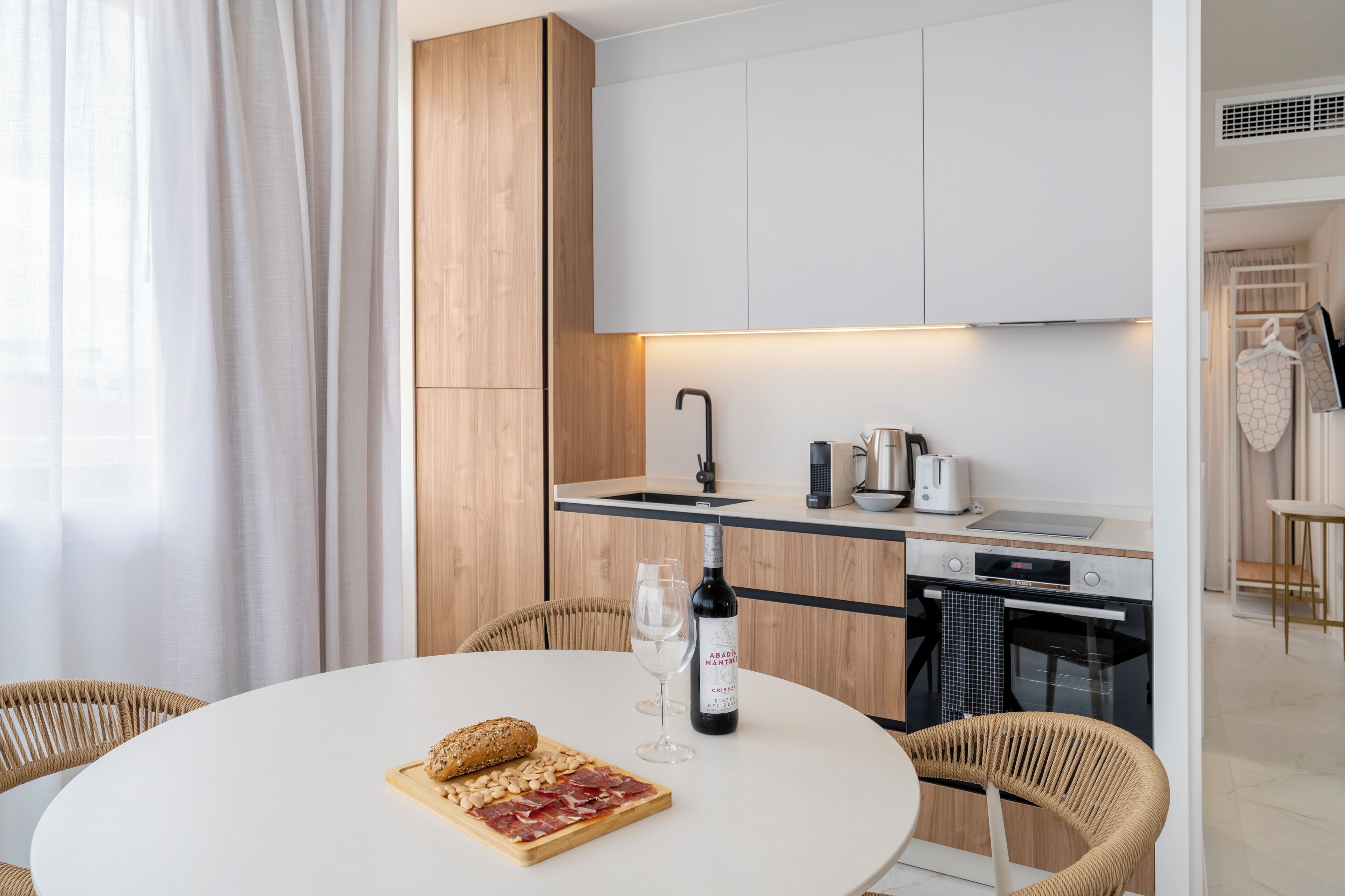 Une cuisine moderne avec des meubles blancs, un robinet noir et un coin repas comprenant une table ronde et des chaises en osier.