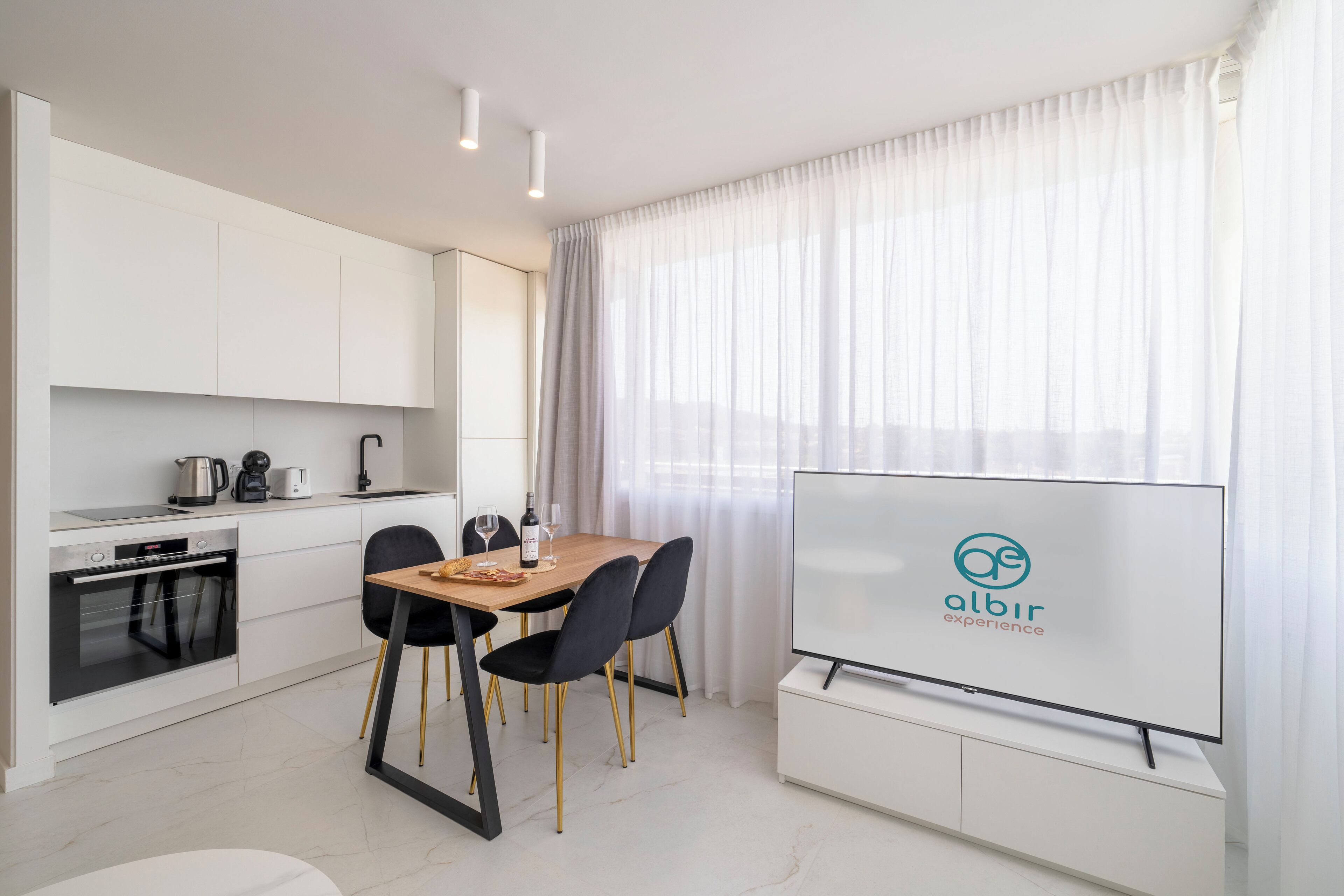 A modern kitchen with white cabinets, a dining table with chairs, a flat-screen TV, and a large window with sheer curtains.