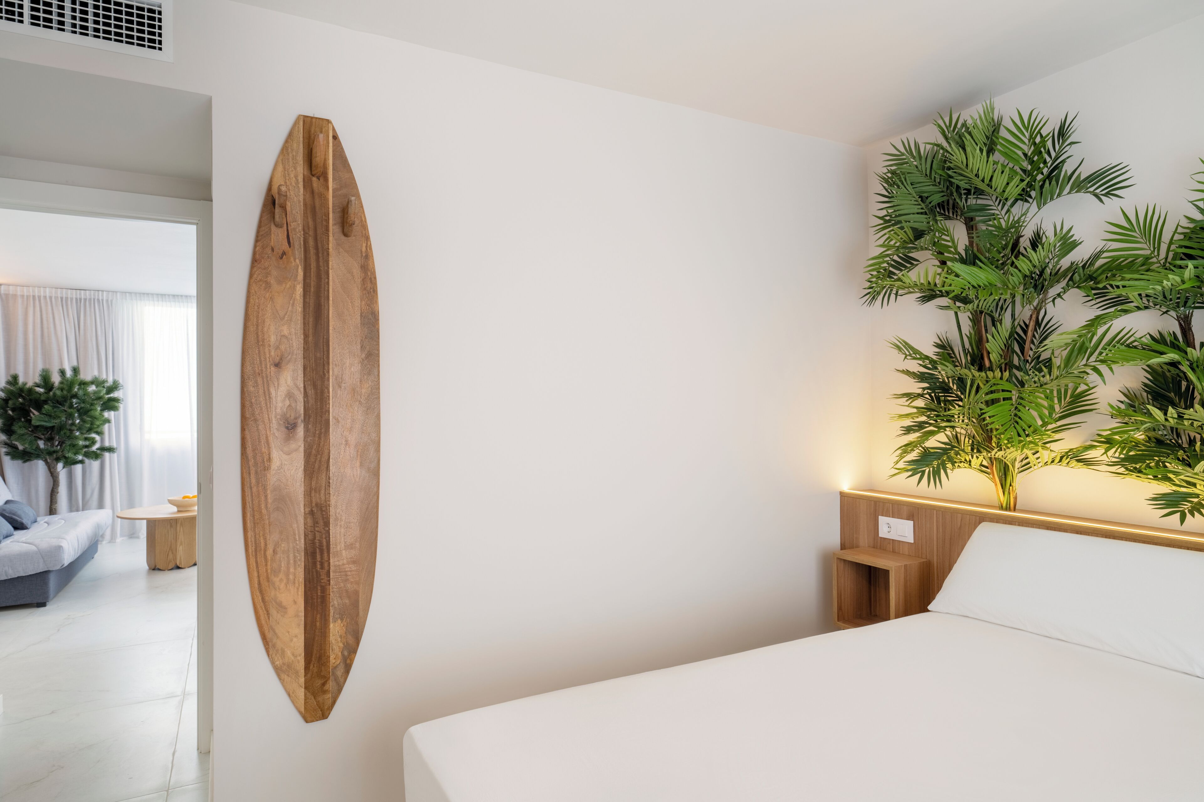 A modern hotel room with a large bed, a wooden surfboard mounted on the wall, a small bedside table, and a potted plant.