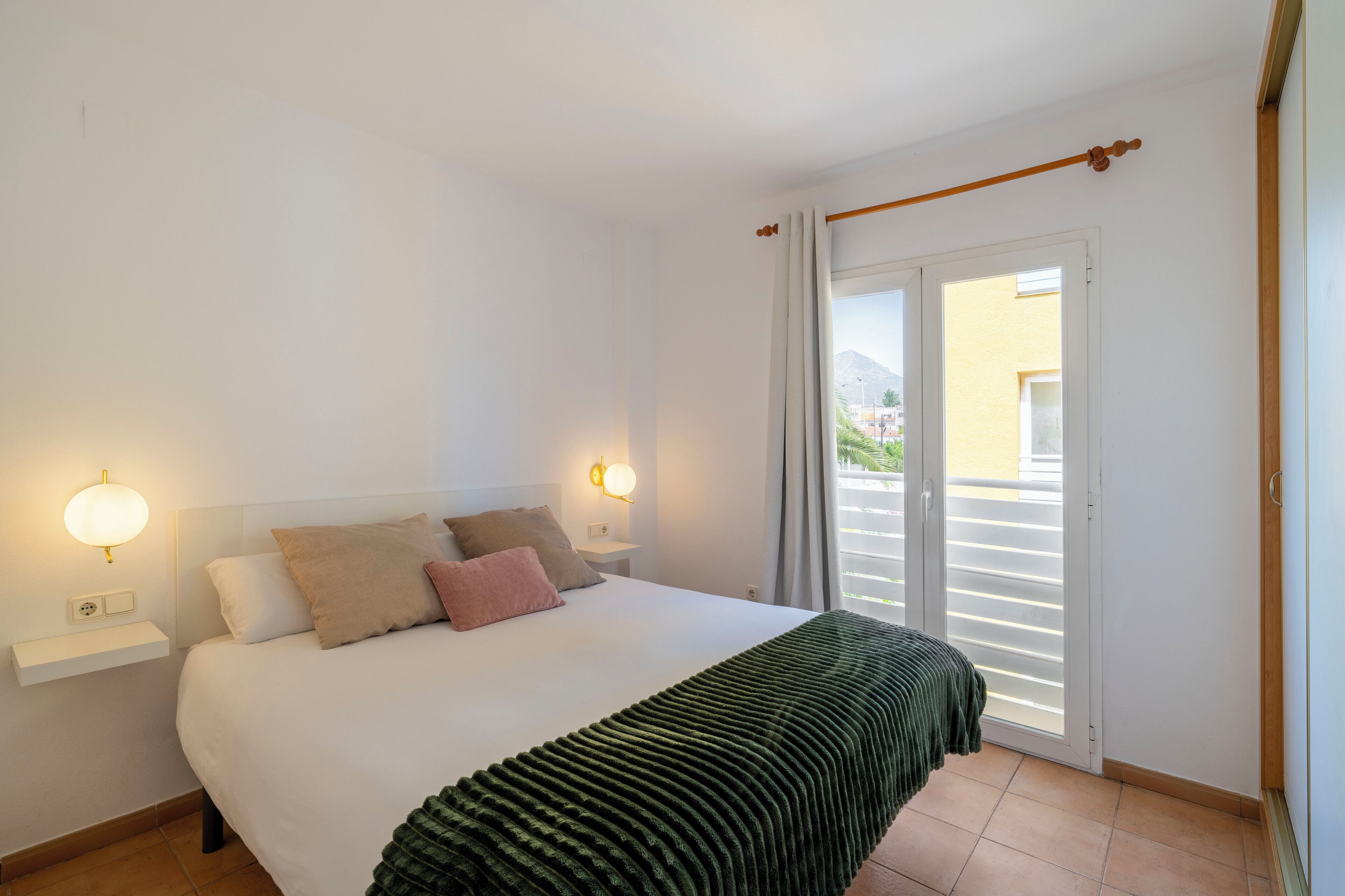 A bedroom with a bed, two bedside lamps, a window with curtains, and a view of the outside.