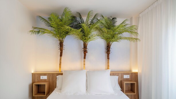 Una habitación de hotel con una cama, dos mesitas de noche y tres palmeras artificiales.