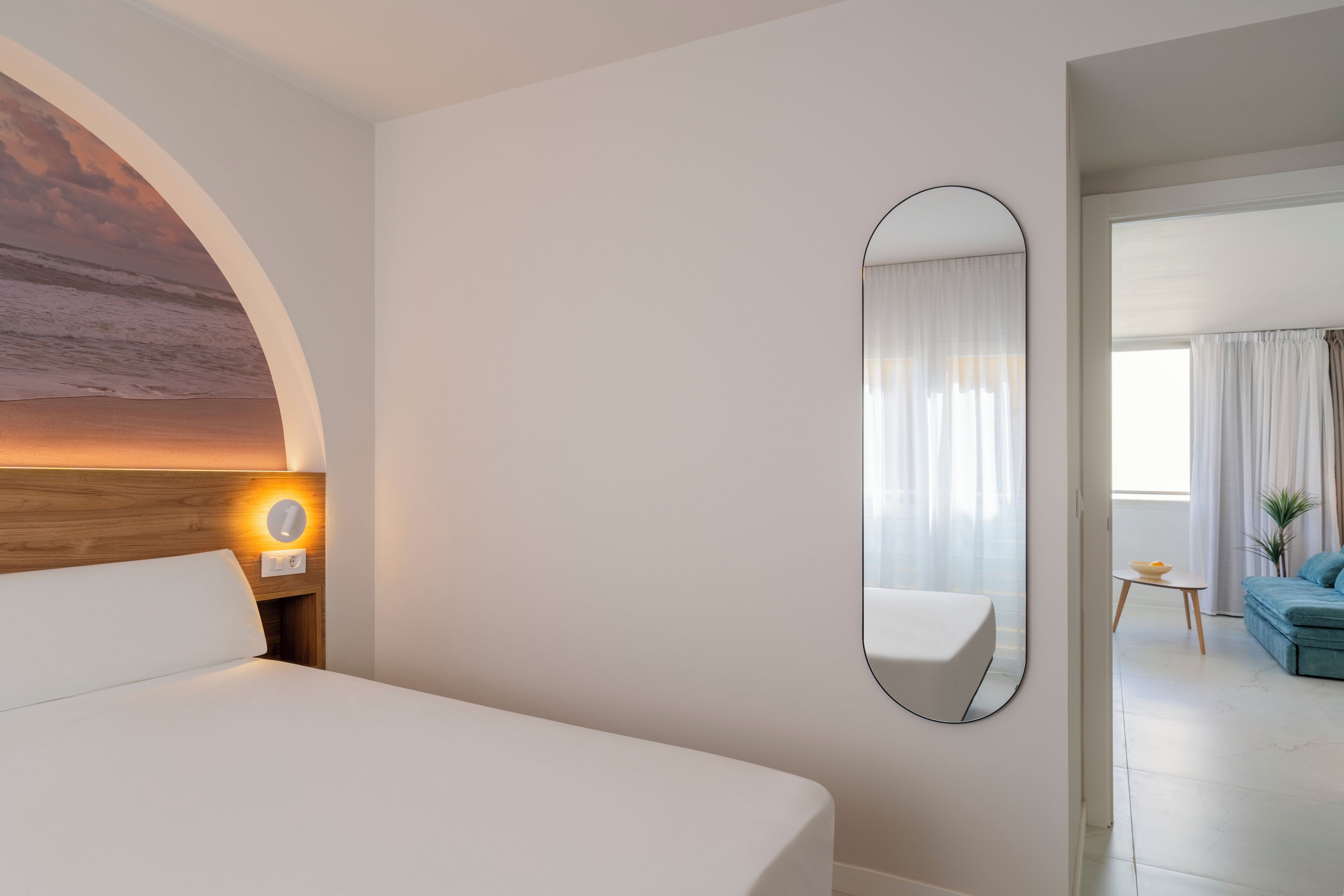 Habitación de hotel con una cama grande, un espejo redondo y vista al área de estar a través de una puerta abierta.