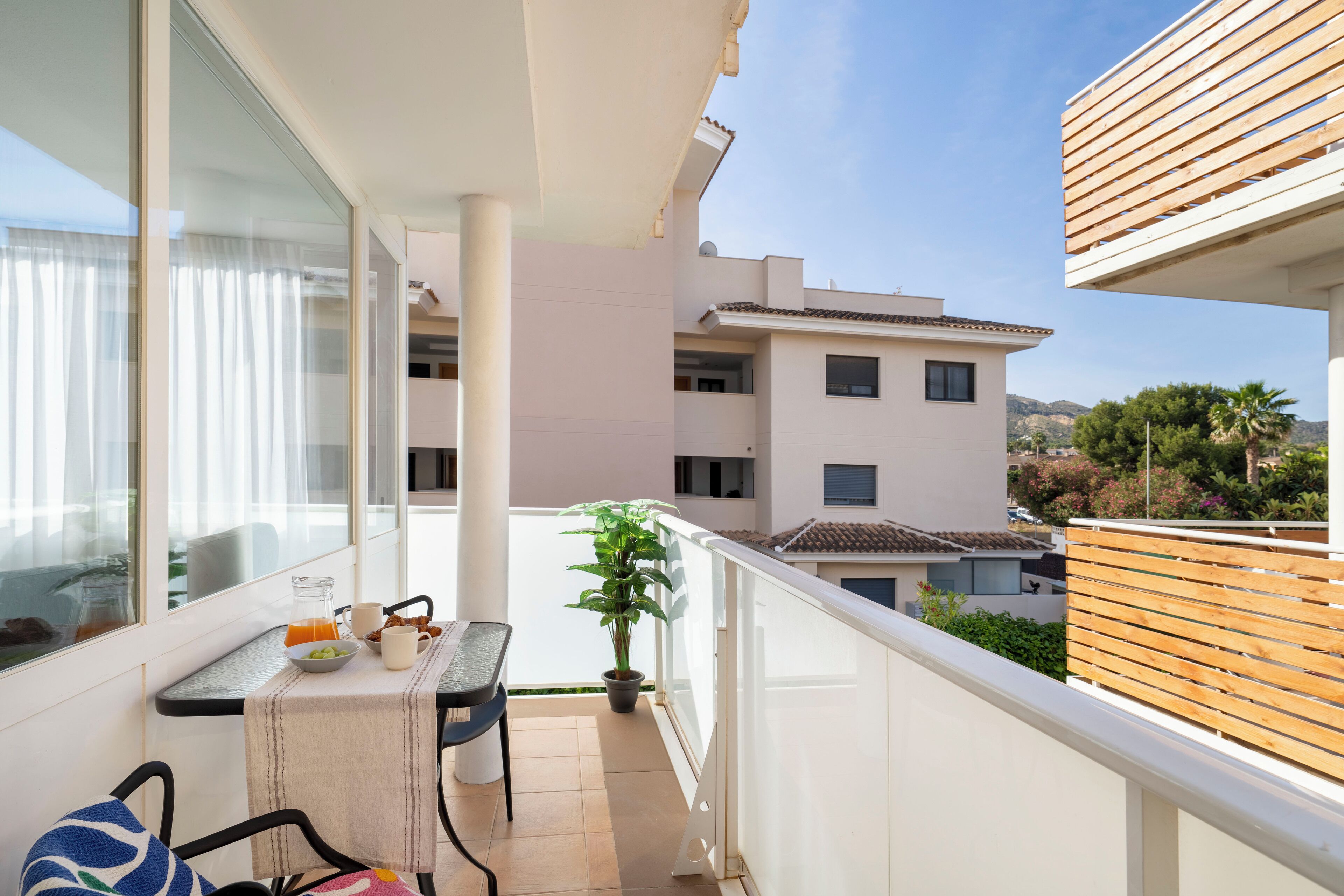 Un balcón con una mesa dispuesta para el desayuno, una planta en maceta y vistas de edificios circundantes.