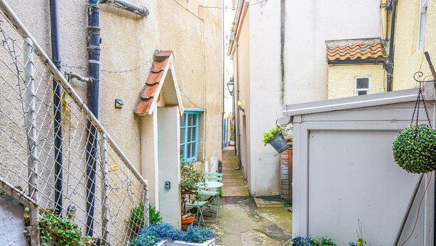 Une ruelle étroite avec une petite table et des chaises, un portail et une plante suspendue.