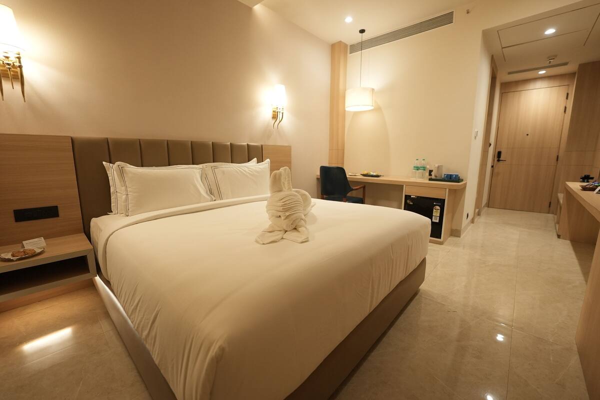 A hotel room with a large bed, a desk with a chair, a minibar, and a bathroom visible in the background.
