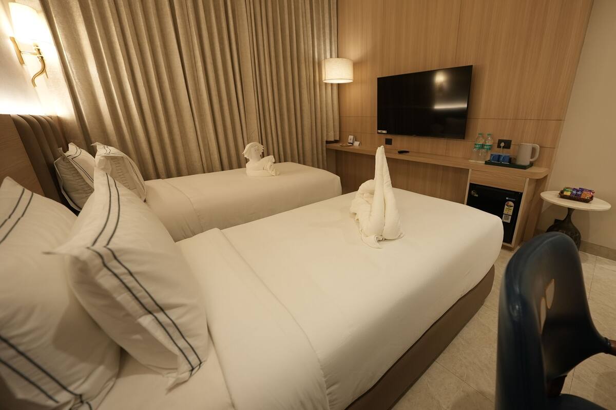 A hotel room with two beds, a flat-screen TV, a minibar, and a small table with a chair.