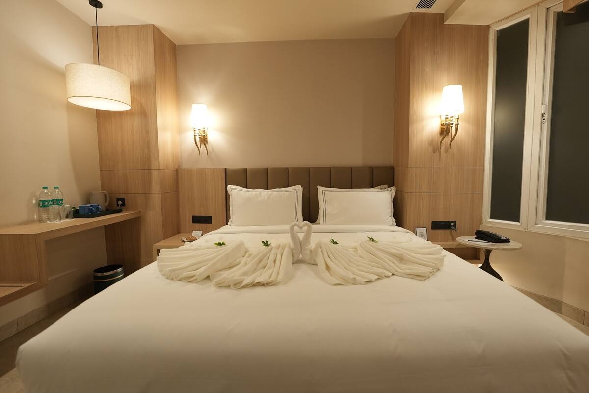 A hotel room with a neatly made bed, two bedside lamps, a bedside table with a phone and a remote control, and a window with blinds.