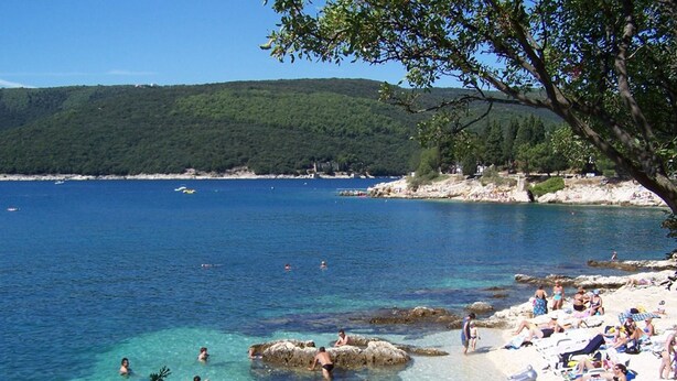 Ein Strand mit klarem Wasser, Felsen und Menschen, die schwimmen und sich sonnen.