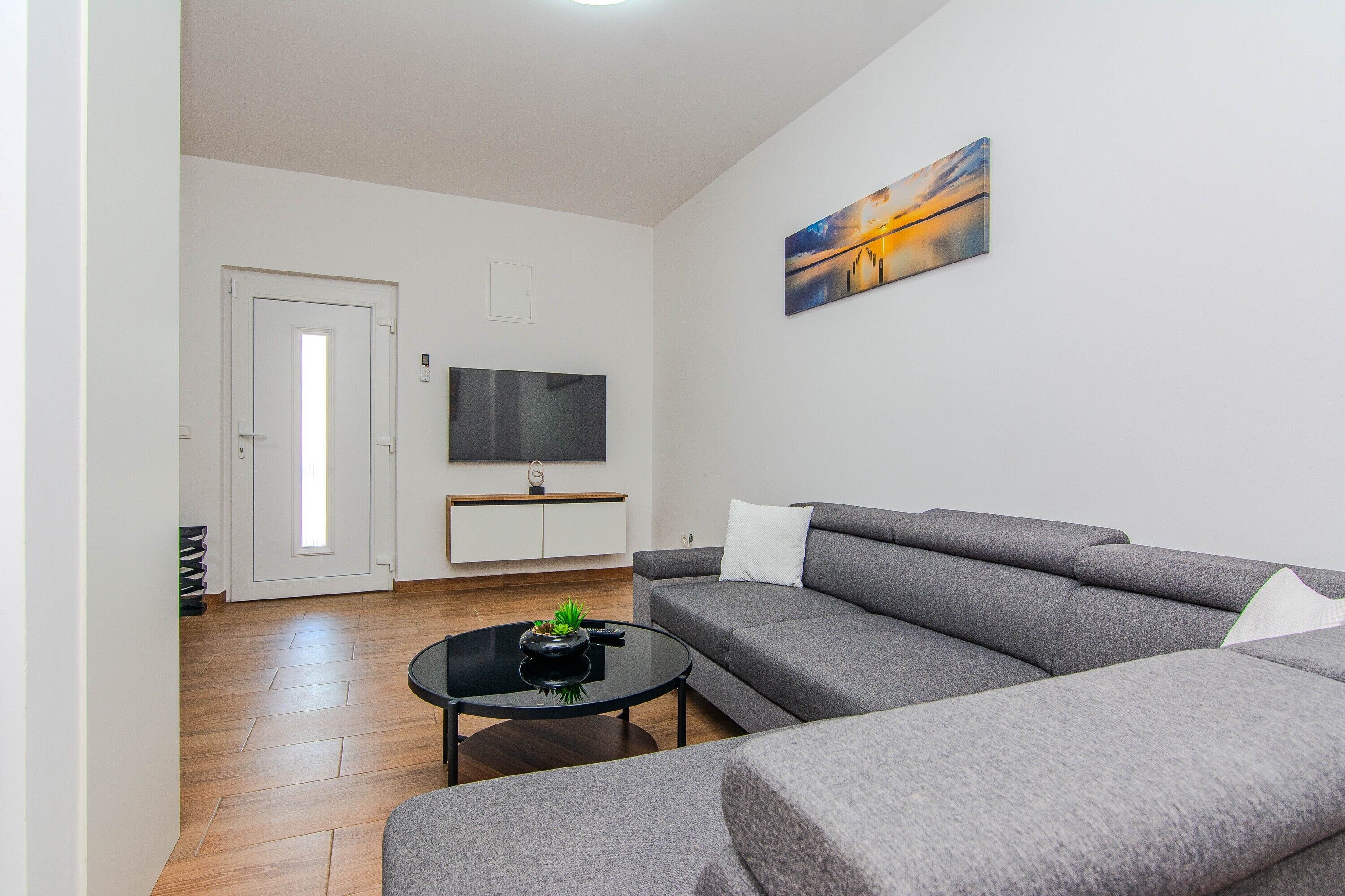 A modern living room with a grey sofa, a black coffee table with a plant, a flat-screen TV mounted on the wall, and a door leading to another room.