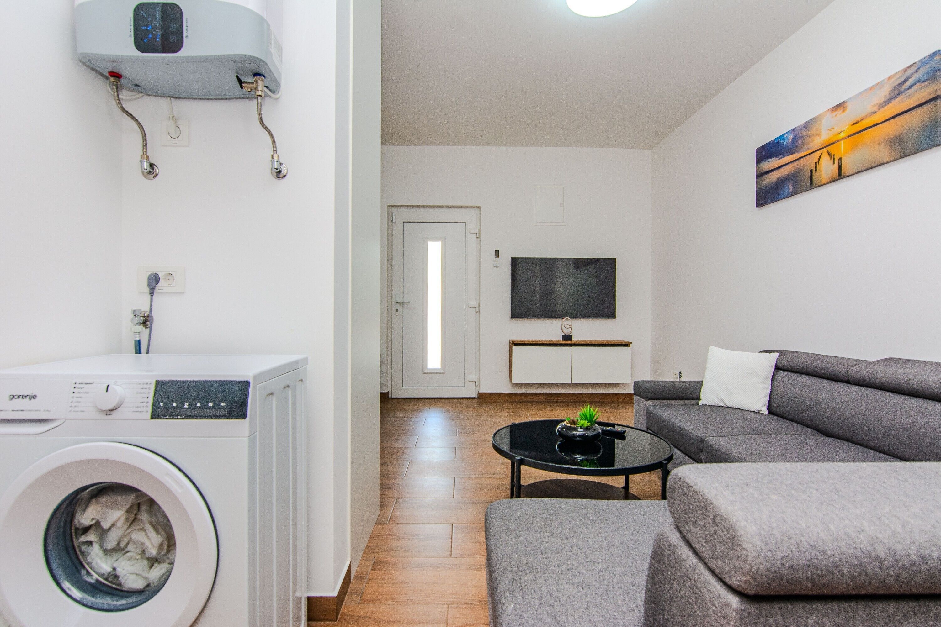 A modern living room with a washing machine, a flat-screen TV, and a grey sofa.