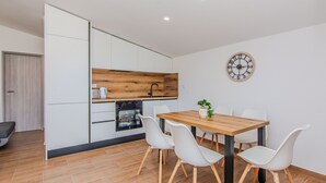 A modern kitchen with white cabinets, a wooden countertop, and a built-in oven. There is a dining table with four white chairs and a clock on the wall. - Apartments Mucaj (Vir)