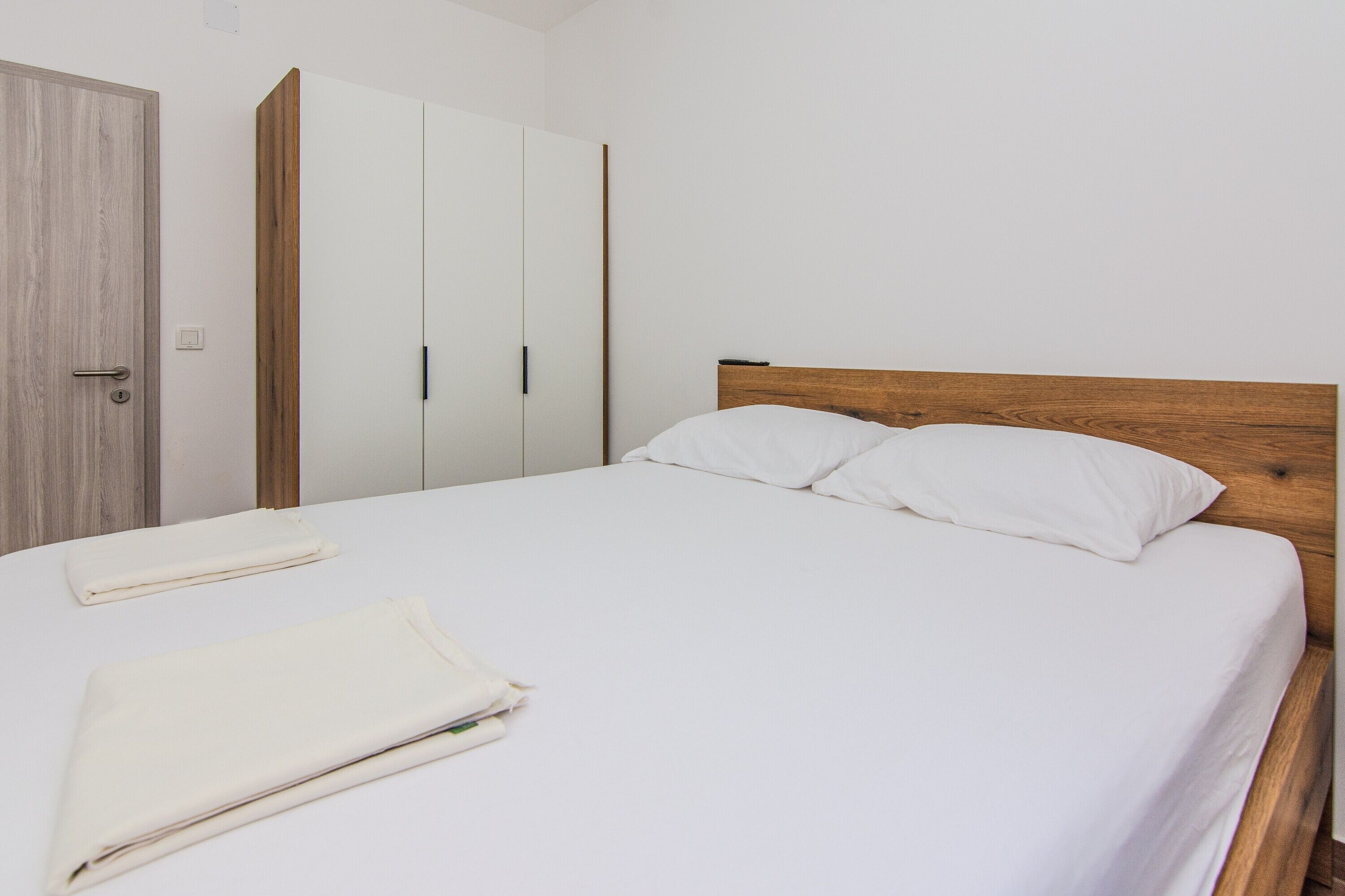 A neatly made bed with white linens and pillows, a wooden headboard, and a white wardrobe with a wooden accent.
