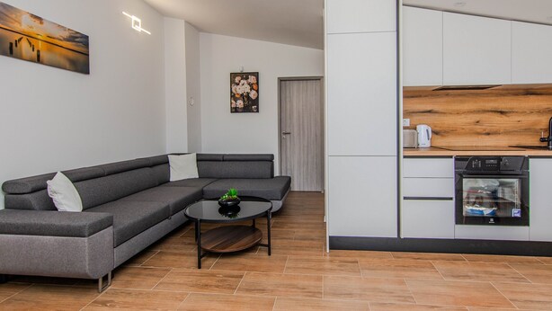 A modern living room with a grey sectional sofa, a round coffee table, and a kitchen area with white cabinets and a wooden countertop. - Apartments Mucaj (Vir)