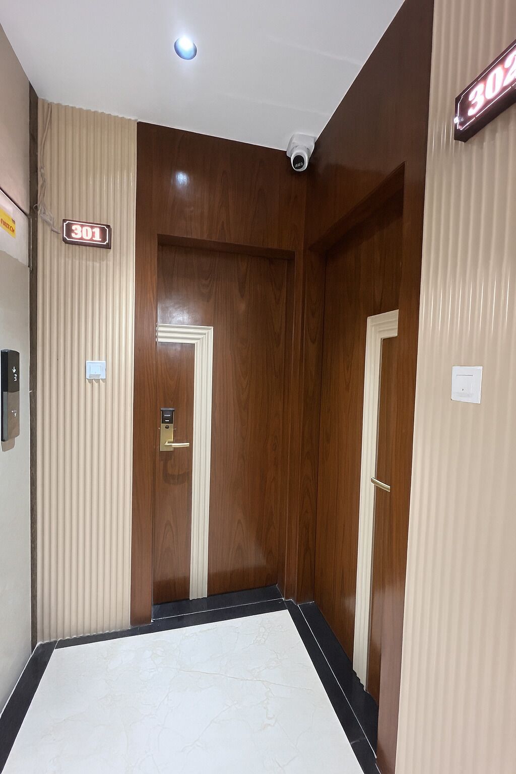 Un couloir avec deux portes en bois, un mur beige orné d’un panneau indiquant un numéro, et un luminaire au plafond.