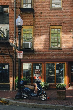 Un edificio de ladrillo con señalización de zona de carga, una moto estacionada al frente y una ventana con un cartel publicitario de un tour por Boston.