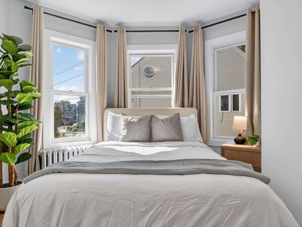 Un dormitorio con cama, cortinas, ventana, radiador y una planta.